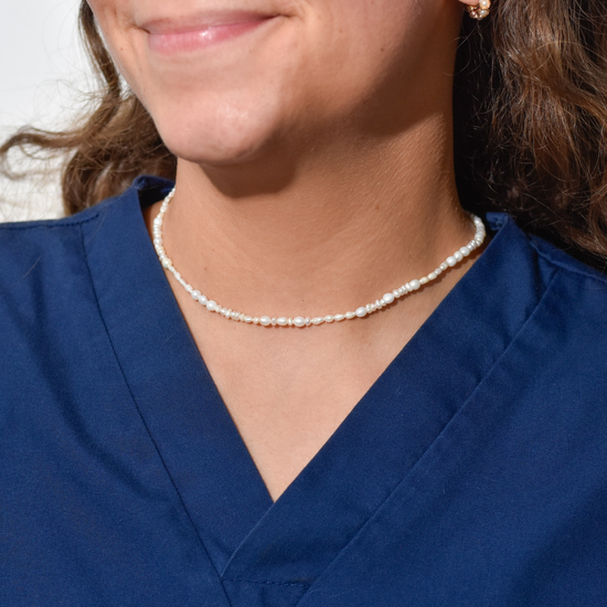 Smiling woman in a blue scrub top wearing a pearl necklace, suggesting a professional, elegant, and confident appearance.