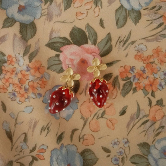 Gold flower earrings with dangling red strawberries adorned with small pearls, set against a pastel floral fabric. Playful and whimsical.