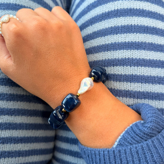 A close-up of a person's wrist wearing a beaded bracelet with dark navy blue polished beads, a white teardrop pearl, and small gold spacer rings, shown against blue and white striped knit clothing.