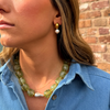 Close-up of a woman wearing a blue denim shirt, a green beaded necklace, and a gold and pearl earring. Background features a brick wall.