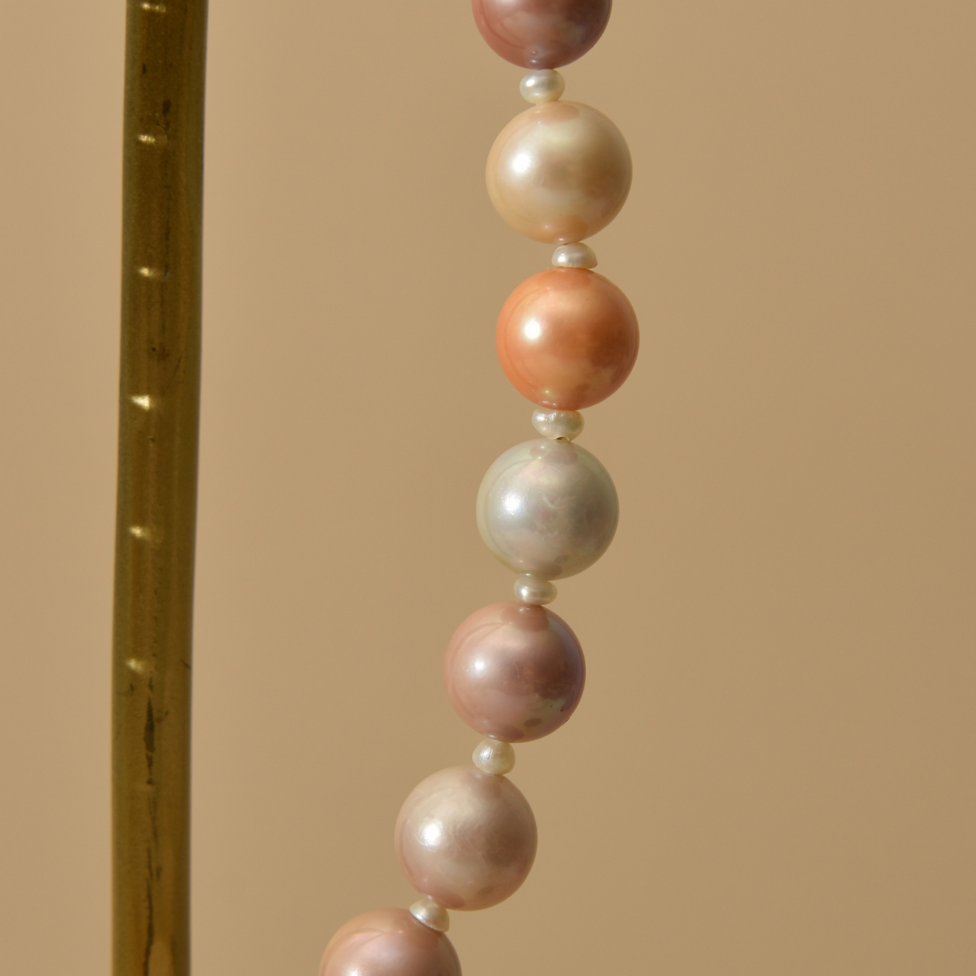 A close-up of large round pearls in soft peach, blush pink, cream, and pale gray tones, separated by small white pearls, against a warm beige background.