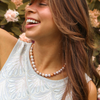 A woman wearing a pearl necklace with a mix of white, cream, and a single peach-colored pearl, paired with a white embroidered sleeveless top against a soft-focus background of green foliage and pink flowers.
