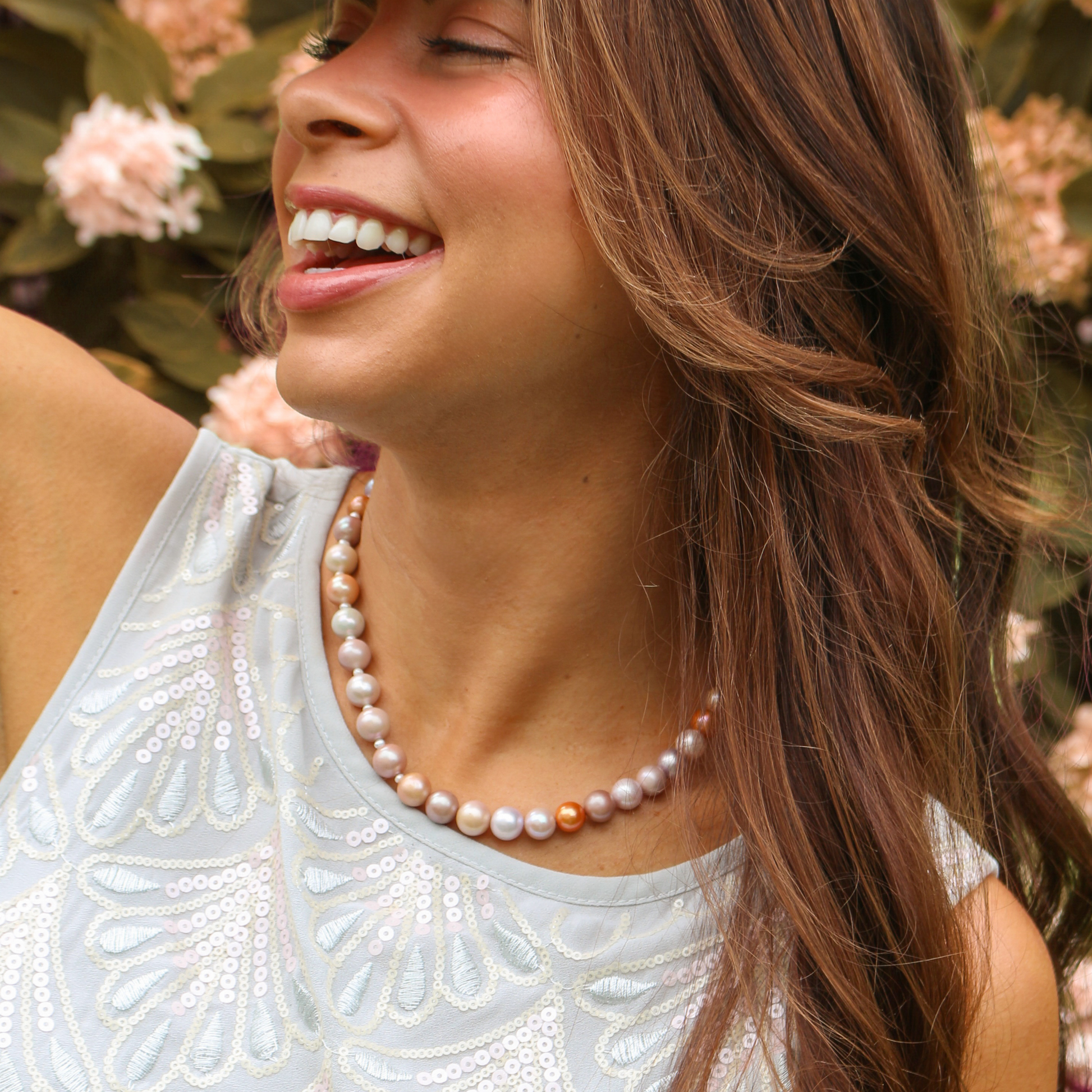 A woman wearing a pearl necklace with a mix of white, cream, and a single peach-colored pearl, paired with a white embroidered sleeveless top against a soft-focus background of green foliage and pink flowers.