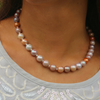 A close-up of a woman's neck and upper chest wearing a strand of freshwater pearls in soft pastel tones including lavender, cream, peach, and taupe, paired with a light gray embroidered top.