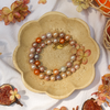 A multicolor freshwater pearl bracelet with orange, gray, and cream pearls, accented by a gold clasp, displayed in a beige flower-shaped ceramic dish on a white fabric background with dried figs and flowers.