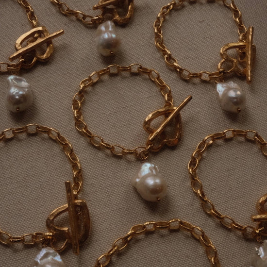 Gold chain bracelets with toggle clasps and irregular pearls are arranged on a textured beige fabric. The tone is elegant and luxurious.
