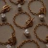 Gold chain bracelets with toggle clasps and irregular pearls are arranged on a textured beige fabric. The tone is elegant and luxurious.
