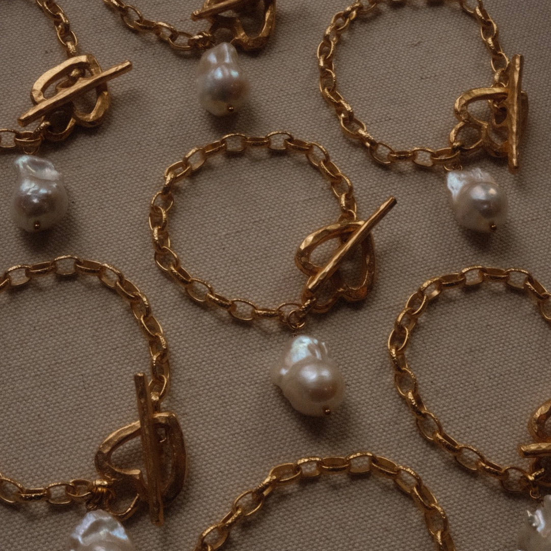 Gold chain bracelets with toggle clasps and irregular pearls are arranged on a textured beige fabric. The tone is elegant and luxurious.