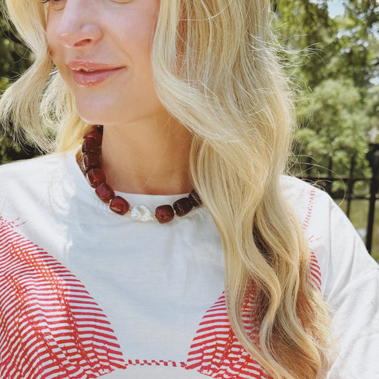 A woman with wavy blonde hair wears a chunky red bead necklace, featuring a large white pearl. She is dressed in a white top with red patterns.