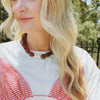 A woman with wavy blonde hair wears a chunky red bead necklace, featuring a large white pearl. She is dressed in a white top with red patterns.