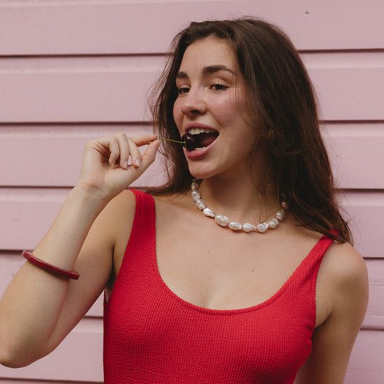 Woman in a red sleeveless top playfully holding a cherry near her mouth, against a pastel pink wall. She wears a pearl necklace and exhibits a cheerful expression.