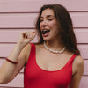 Woman in a red sleeveless top playfully holding a cherry near her mouth, against a pastel pink wall. She wears a pearl necklace and exhibits a cheerful expression.