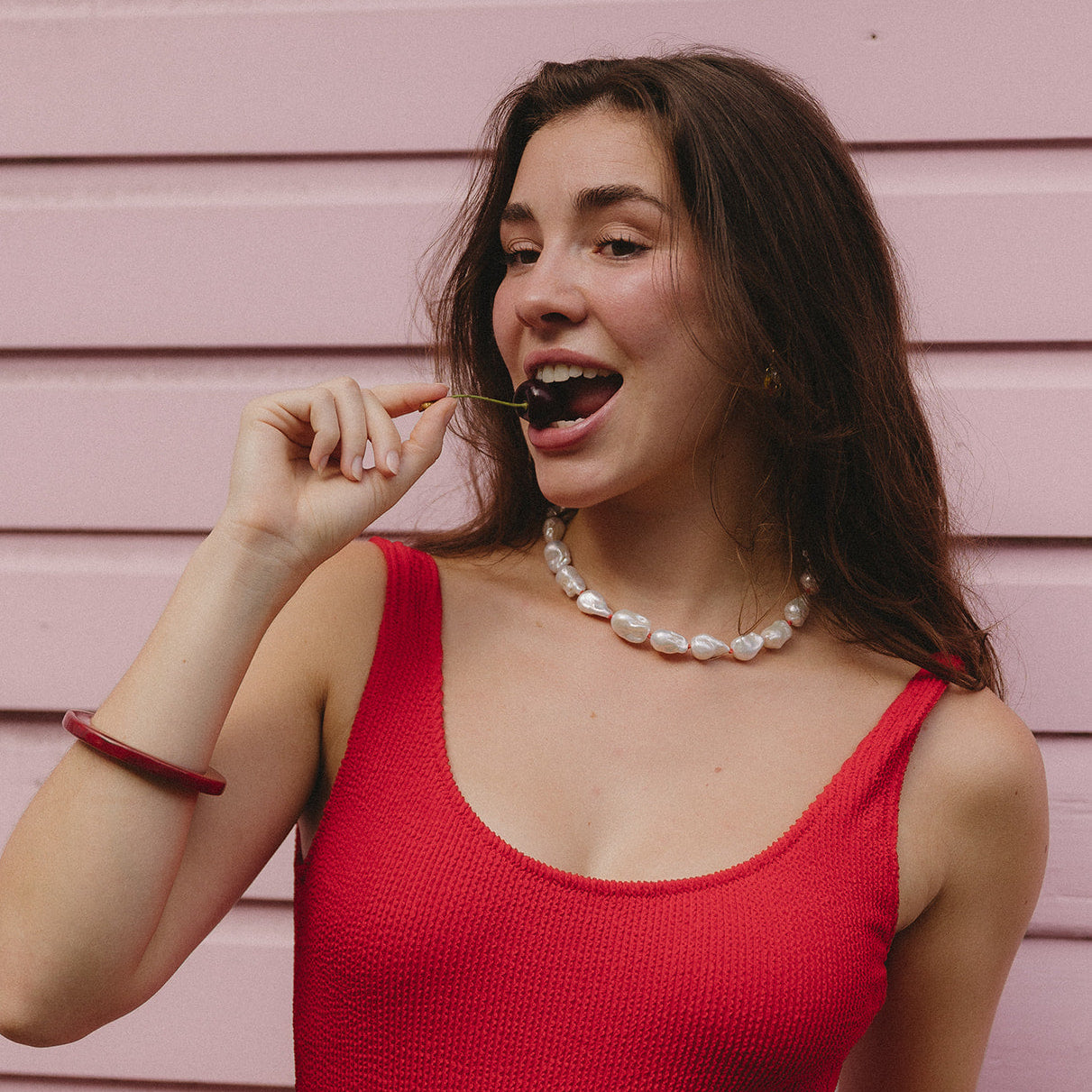 Woman in a red sleeveless top playfully holding a cherry near her mouth, against a pastel pink wall. She wears a pearl necklace and exhibits a cheerful expression.
