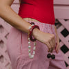Close-up of a person in a red top and pink striped pants, holding cherries. They have a pearl and red bangle bracelet, set against a pink wooden background.