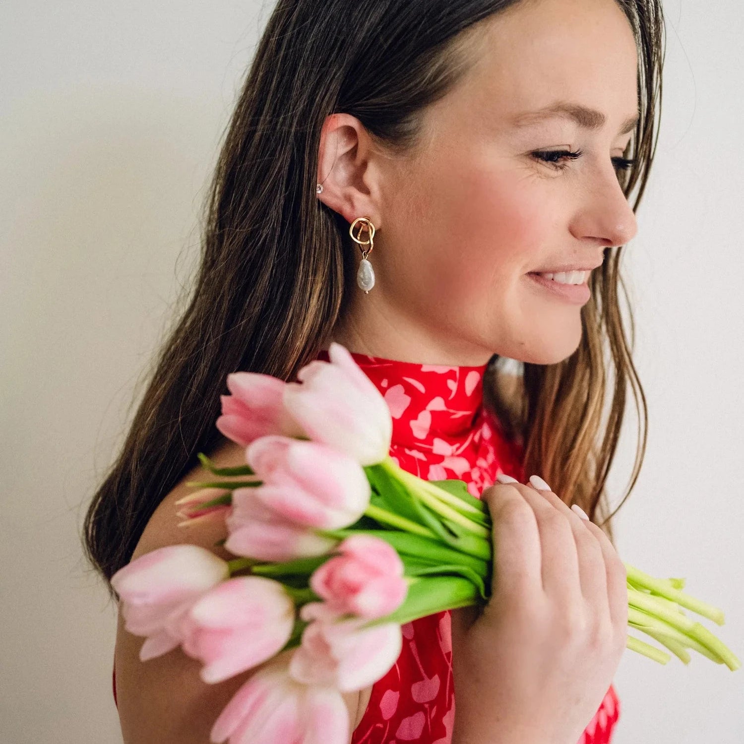 A woman in a red heart-patterned dress holds pink tulips, smiling softly. She wears gold and pearl earrings, conveying a warm, joyful tone.