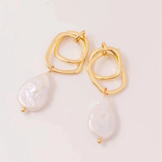 Gold earrings with intertwined geometric loops and dangling baroque pearl drops on a light background, conveying elegance and sophistication.