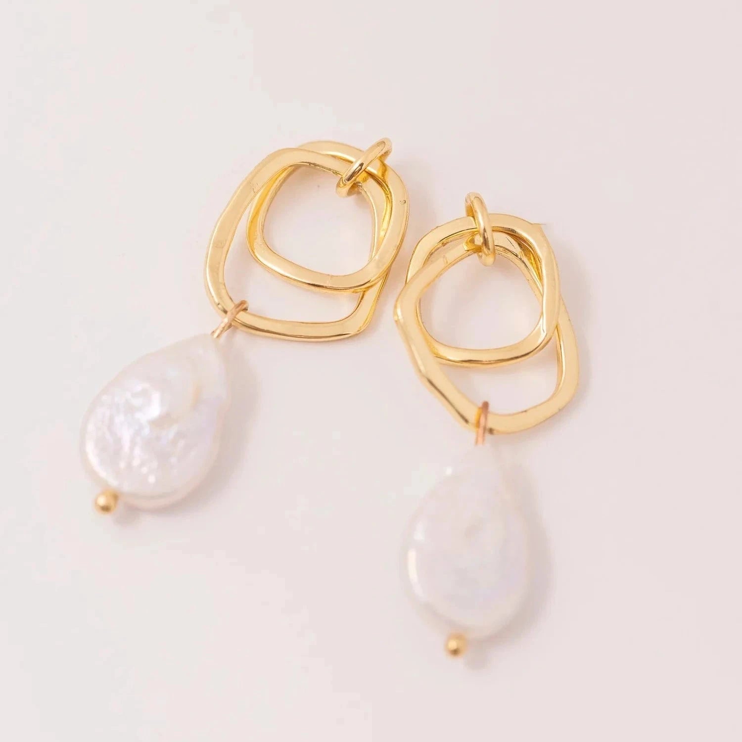 Gold earrings with intertwined geometric loops and dangling baroque pearl drops on a light background, conveying elegance and sophistication.