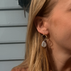 Close-up of a woman’s ear with blonde hair, wearing a gold earring featuring a geometric shape and a dangling white pearl, against a soft background.