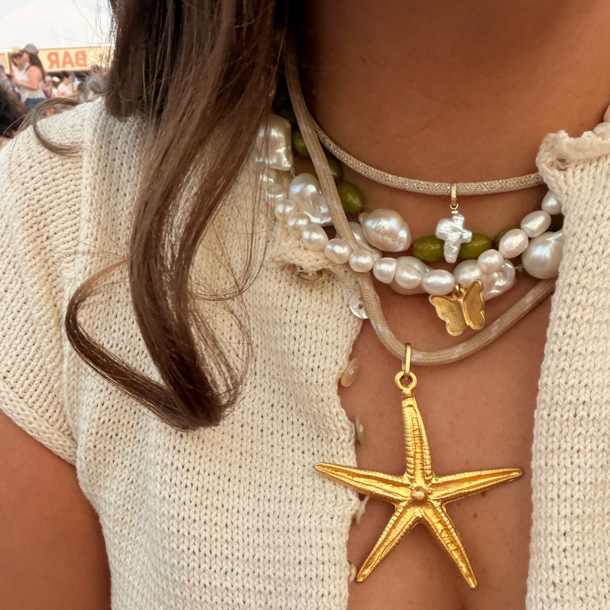 A close-up of a person wearing a layered necklace set. The necklace features a large gold starfish pendant, a gold butterfly charm, a white pearl strand, and green beads on a beige cord, worn over a cream knit top.