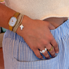 A close-up of a person's hand resting in the pocket of blue and white striped pants, showcasing gold rings, a gold watch, and stacked bracelets, including a cross charm.