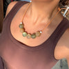 A close-up of a person wearing a beaded necklace with matte olive-green and beige round beads on an orange cord, paired with a brown ribbed tank top.
