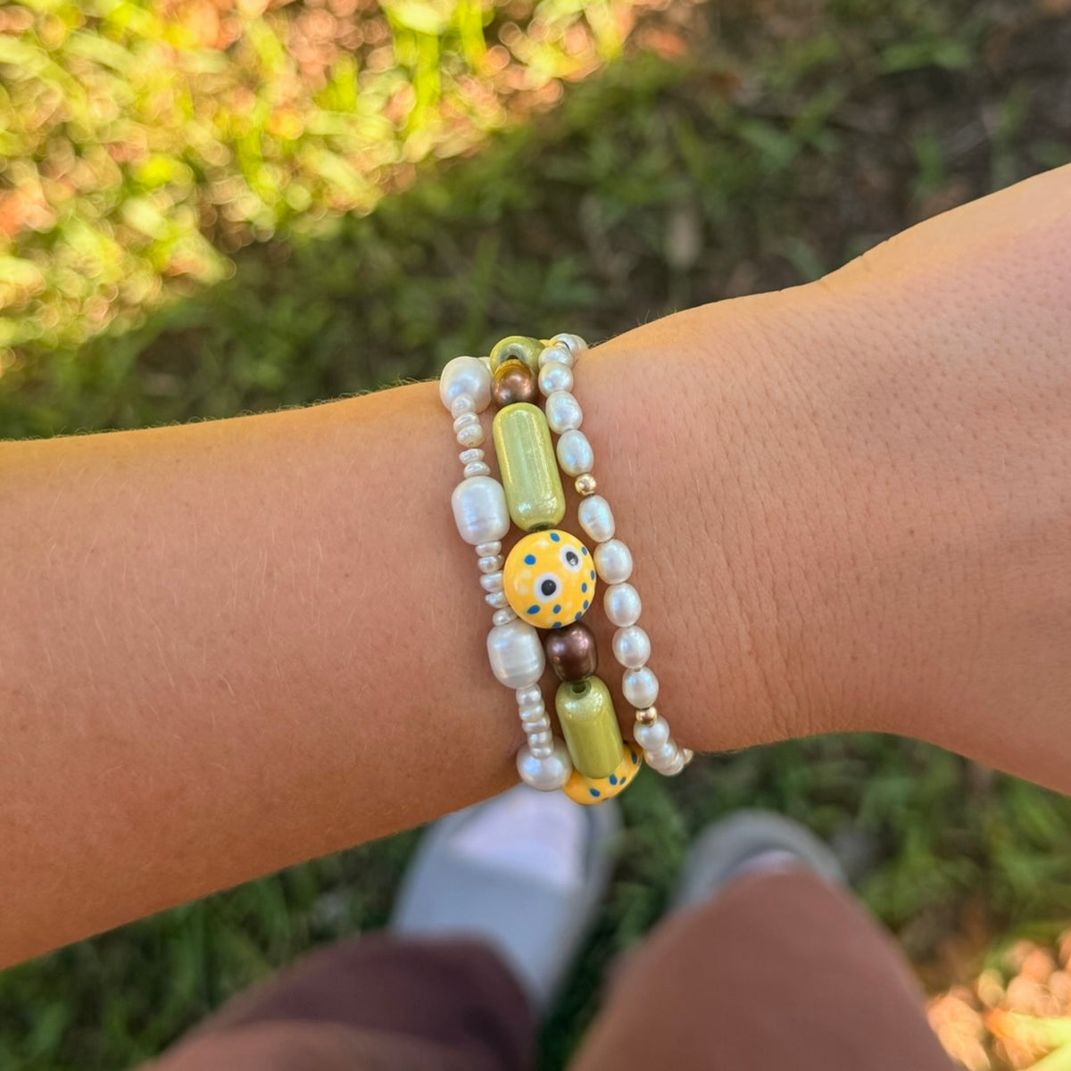 A close-up of a person's wrist wearing a stack of three beaded bracelets outdoors on a grassy background. The bracelets include white faux pearls, olive green and yellow-green cylindrical beads, a yellow bead with blue and black dots, and a brown bead.