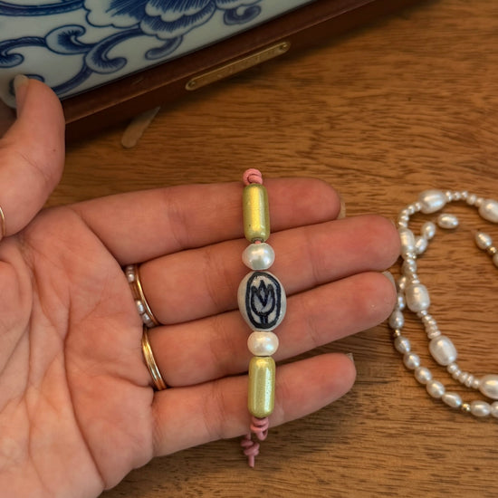 A handmade beaded bracelet held in a person's hand, featuring pale yellow-green beads, white pearls, and a central oval bead with a dark blue floral design. A matching pearl necklace is visible on the wooden table background.