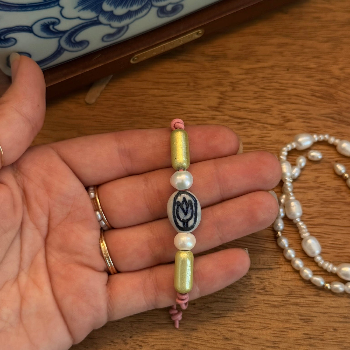 A handmade beaded bracelet held in a person's hand, featuring pale yellow-green beads, white pearls, and a central oval bead with a dark blue floral design. A matching pearl necklace is visible on the wooden table background.