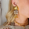 A close-up of an ear with a unique earring featuring a gold fish head and tail with the body as pearls. The person wears a woven top and a gold necklace.