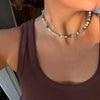 A close-up of a person wearing a beaded choker necklace with white pearls and small green and iridescent beads, paired with a brown ribbed tank top.