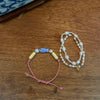 Two handmade beaded jewelry pieces displayed on a wooden tabletop: a pink cord bracelet with pale yellow and blue beads, and a multi-strand pearl necklace with a small gold leaf charm.
