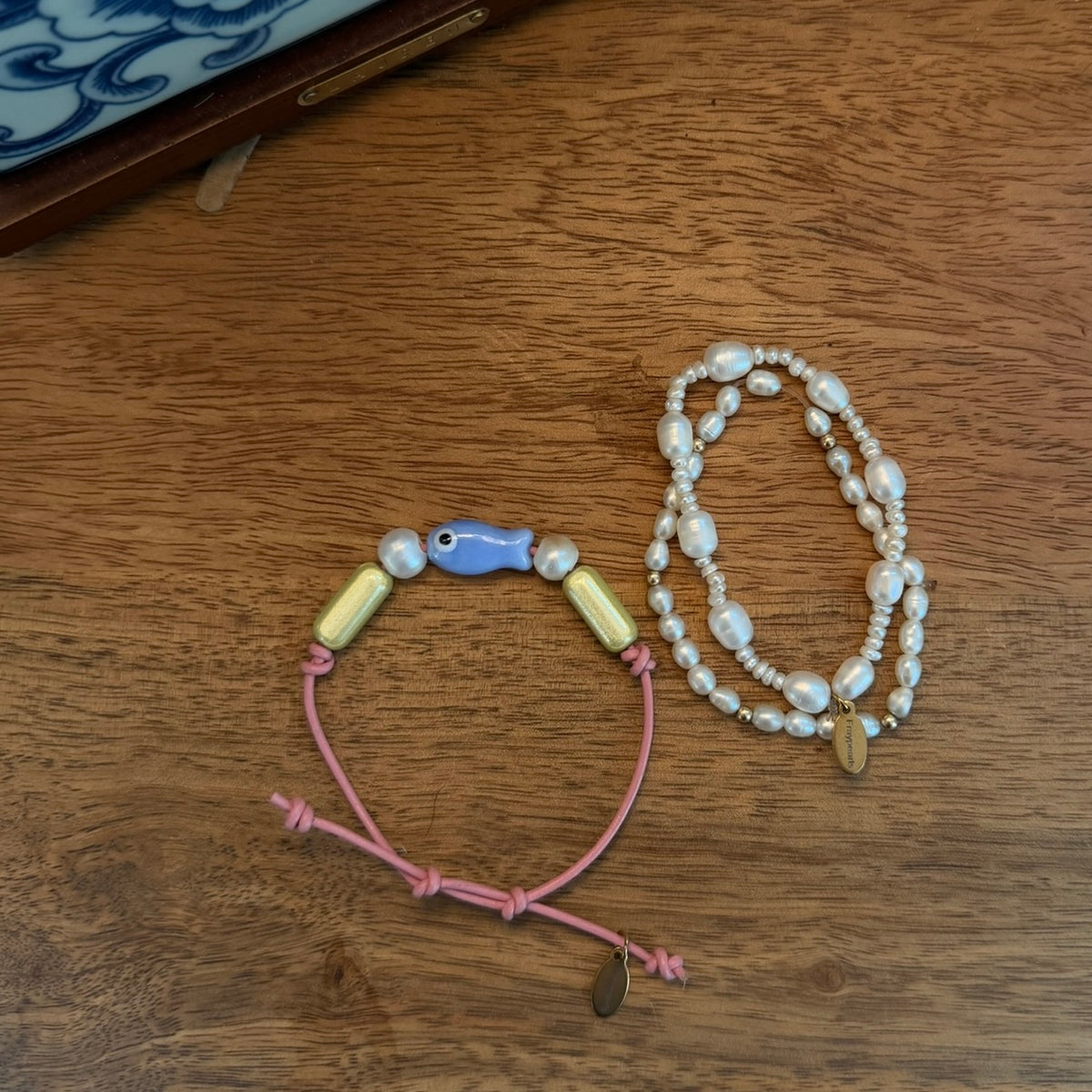 Two handmade beaded jewelry pieces displayed on a wooden tabletop: a pink cord bracelet with pale yellow and blue beads, and a multi-strand pearl necklace with a small gold leaf charm.