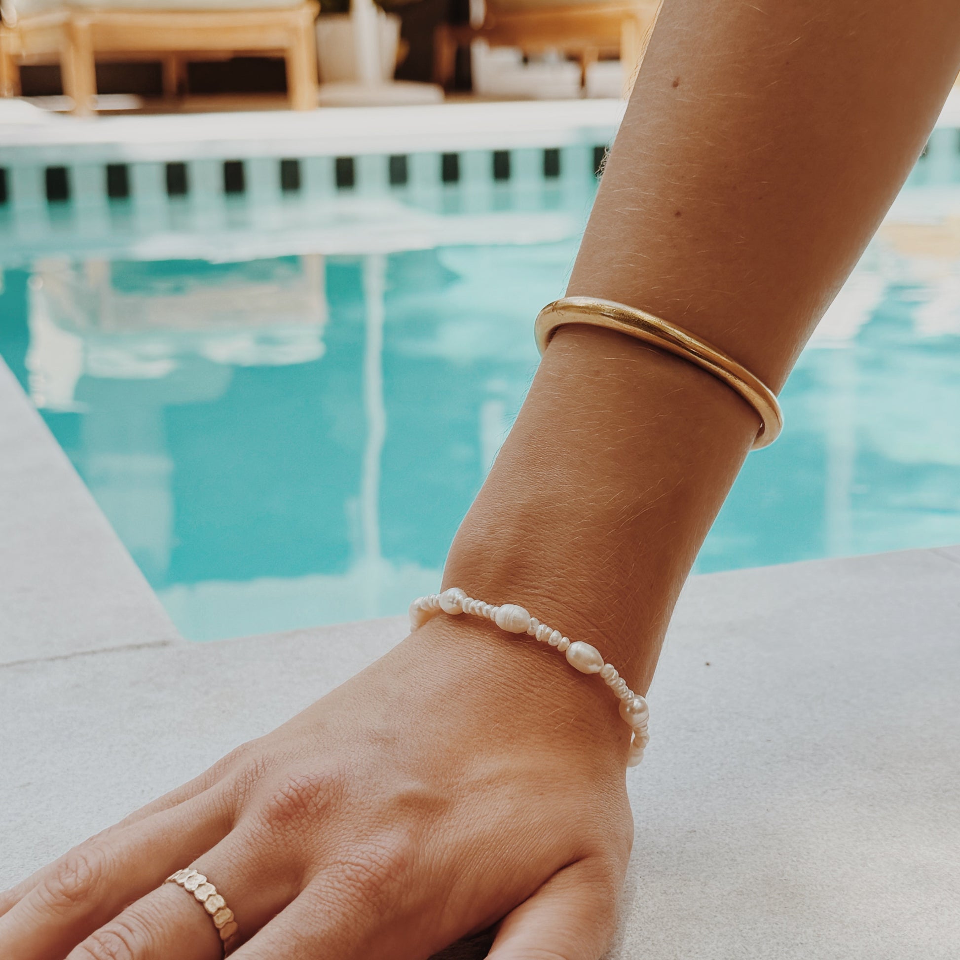 Close-up of a hand on poolside, wearing a gold bangle, pearl bracelet, and ring. The setting conveys a relaxed, luxurious summer vibe.