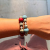 A close-up of a person's wrist wearing two beaded bracelets. One bracelet features smooth red and light blue beads, while the other features speckled cream and light blue beads.