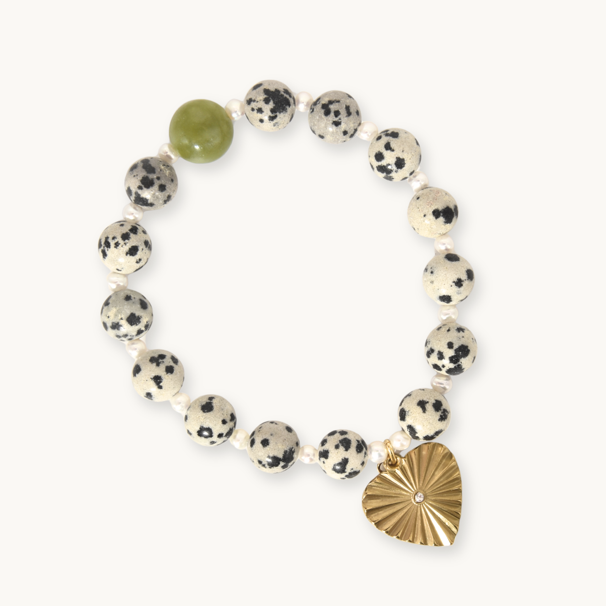 A beaded stretch bracelet featuring speckled cream and black stone beads, a single smooth olive green bead, and a gold-toned sunburst heart charm with a small clear rhinestone accent, shown on a light off-white background.
