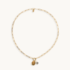 A delicate gold paperclip chain necklace with two small hanging charms, one gold-toned and one jasper and pear, displayed on a clean off-white background.