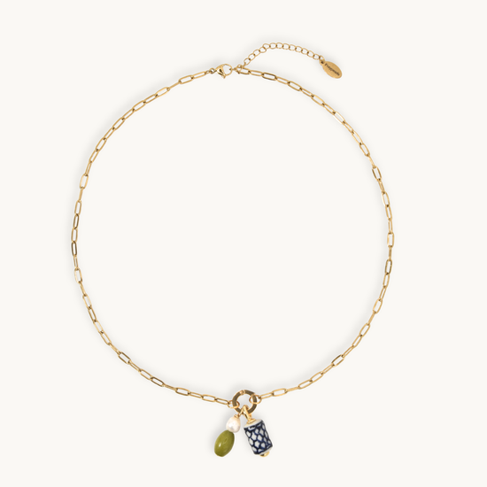 A gold chain necklace with a small ring charm holding an olive green bead and a navy blue patterned pouch, displayed on a clean off-white background.