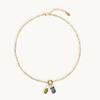 A gold chain necklace with a small ring charm holding an olive green bead and a navy blue patterned pouch, displayed on a clean off-white background.