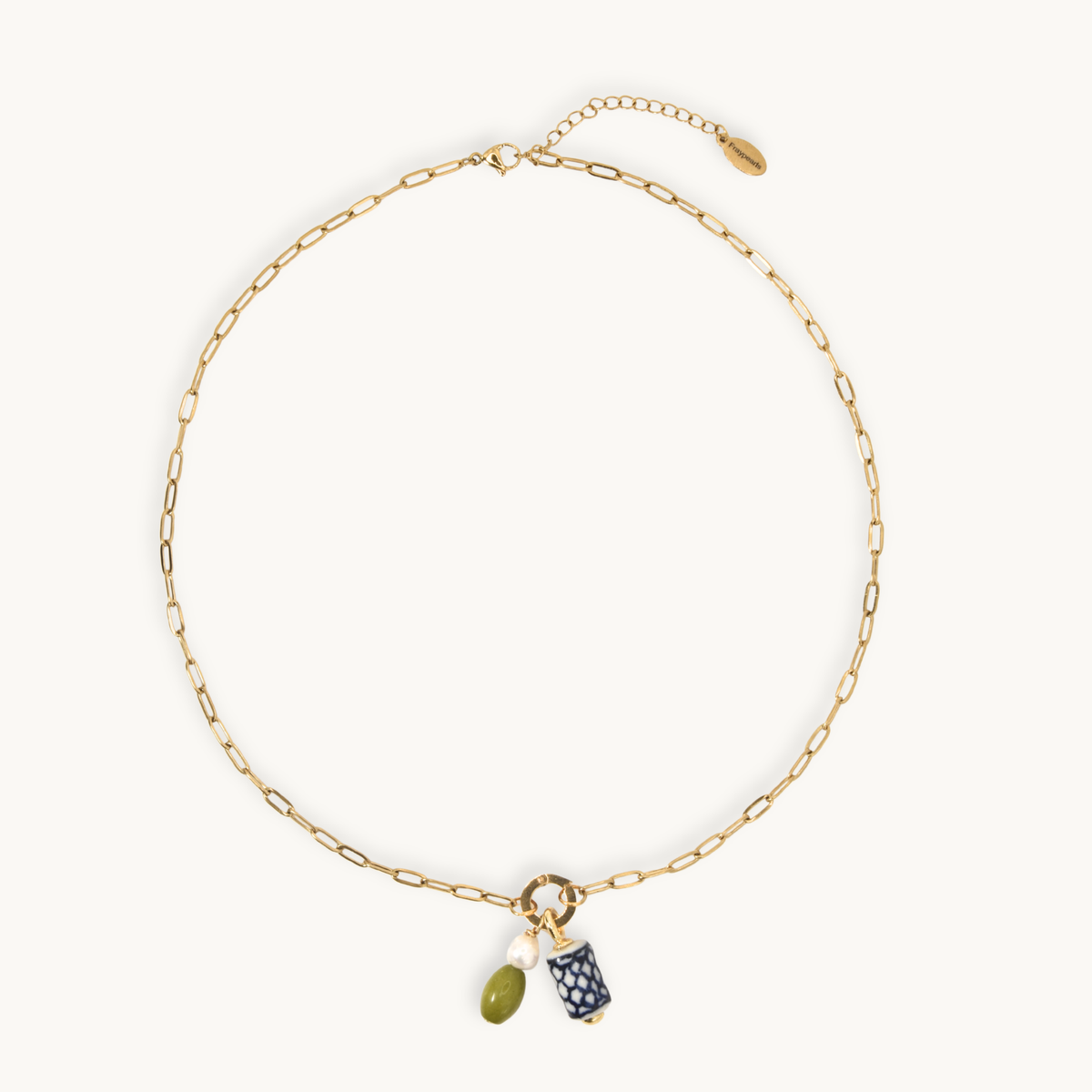A gold chain necklace with a small ring charm holding an olive green bead and a navy blue patterned pouch, displayed on a clean off-white background.