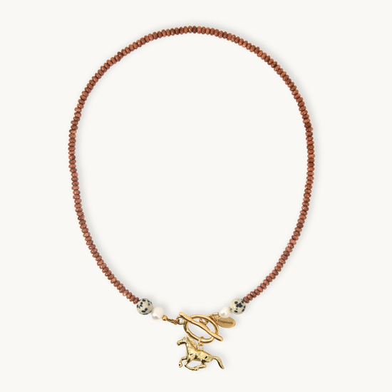A delicate necklace featuring a strand of small glittery orange beads, accented with white beads, a gold toggle clasp, and a gold horse charm, displayed on a clean off-white background.