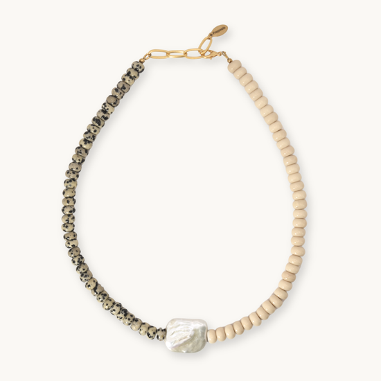 A beaded necklace featuring a textured black-and-white speckled strand, a strand of light beige beads, a white baroque pearl, and a gold-tone chain clasp, shown on a light cream background.