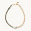 A beaded necklace featuring a textured black-and-white speckled strand, a strand of light beige beads, a white baroque pearl, and a gold-tone chain clasp, shown on a light cream background.