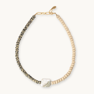 A beaded necklace featuring a textured black-and-white speckled strand, a strand of light beige beads, a white baroque pearl, and a gold-tone chain clasp, shown on a light cream background.