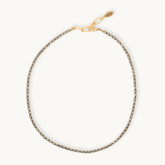 A delicate smoky brown rice pearls with a golden chain and closure necklace with a small oval tag, displayed in a neat loop on a clean off-white background.
