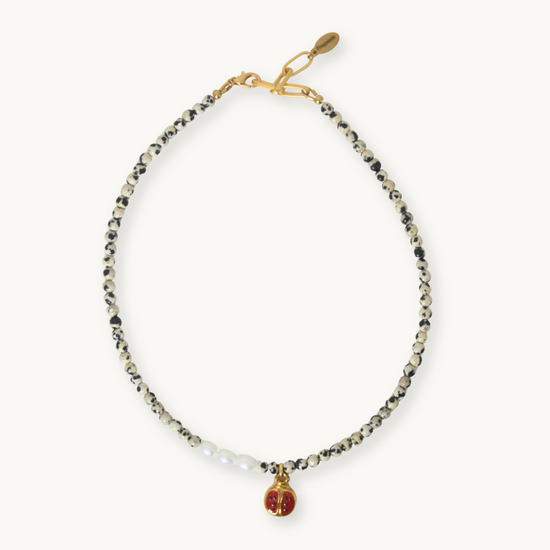 A delicate beaded necklace with black and white beads, a small cluster of white pearls, and a gold-tone charm featuring a red enamel heart with a gold 'F' monogram, displayed on a light cream background.