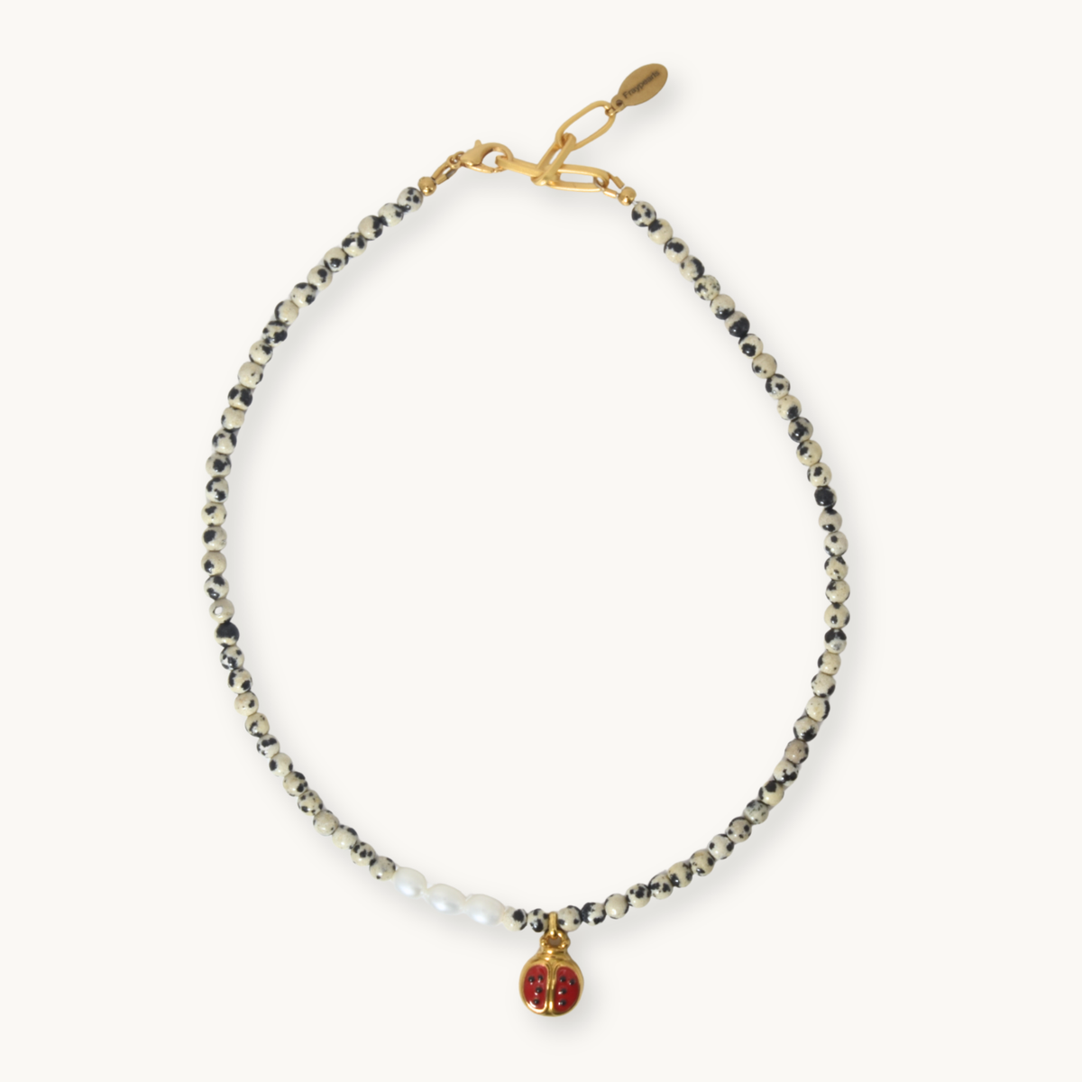 A delicate beaded necklace with black and white beads, a small cluster of white pearls, and a gold-tone charm featuring a red enamel heart with a gold 'F' monogram, displayed on a light cream background.