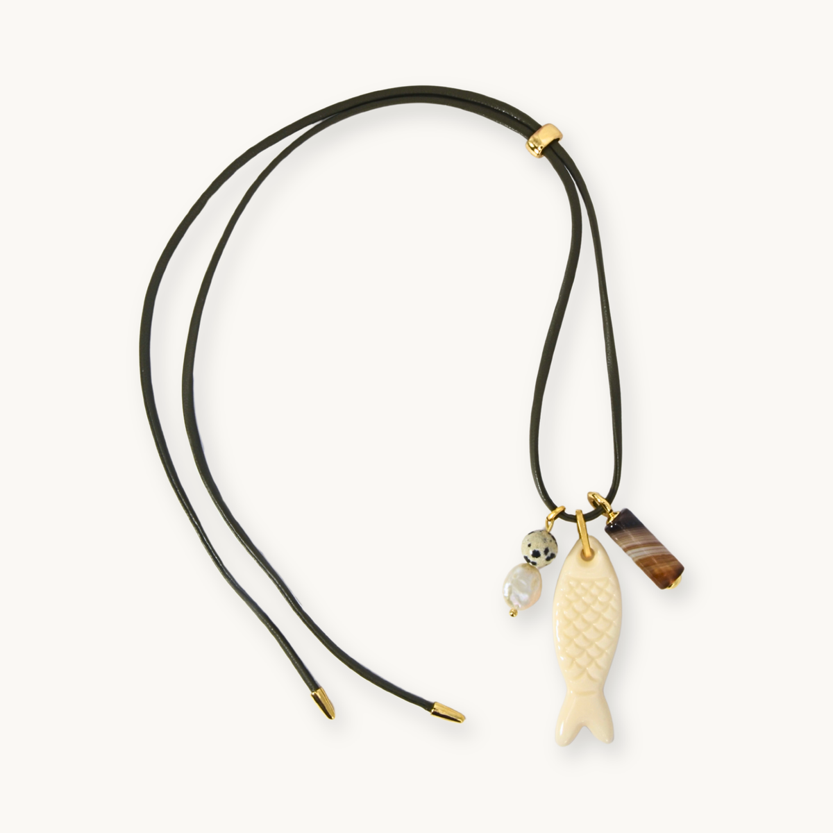 A double-strand black cord necklace with gold-toned hardware, featuring a carved ivory fish pendant, a small white bead charm, and a brown rectangular stone charm, displayed on a clean off-white background.