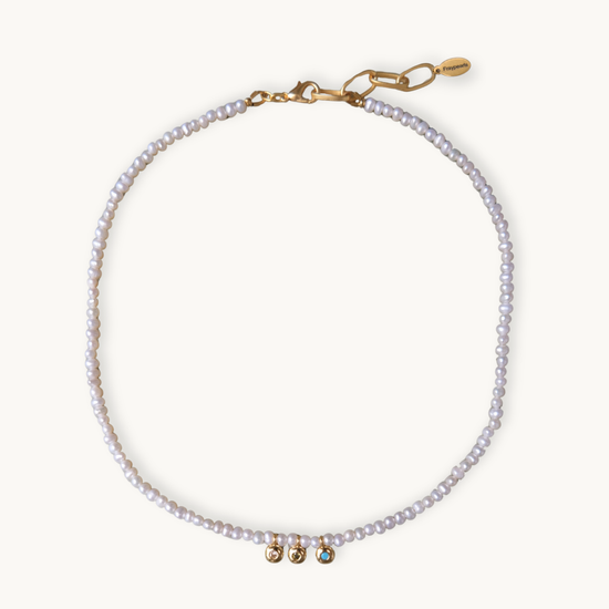 A delicate pearl necklace with small white pearls and gold-tone hardware, featuring three small decorative beads at the center and a lobster clasp closure, shown on a light cream background.