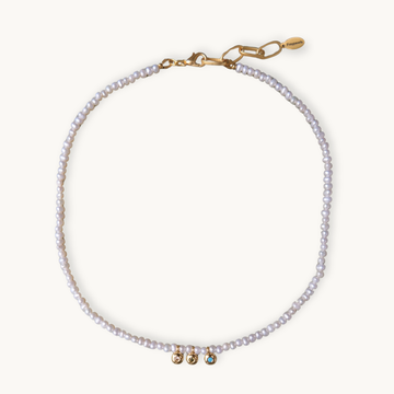 A delicate pearl necklace with small white pearls and gold-tone hardware, featuring three small decorative beads at the center and a lobster clasp closure, shown on a light cream background.