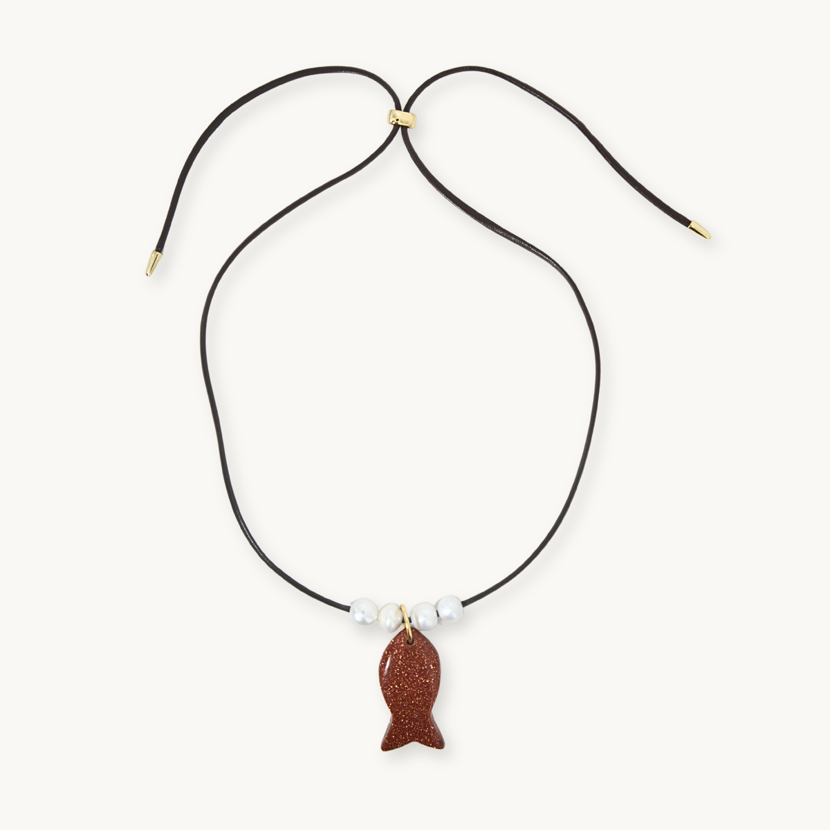 A black cord necklace with a small brown textured fish pendant, accented by three white beads and gold-tone hardware, displayed on a light cream background.